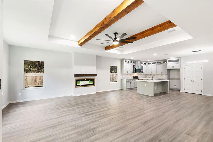 Unfurnished living room featuring a raised ceiling, a multi sided fireplace, light wood-type flooring, a ceiling fan, and beamed ceiling