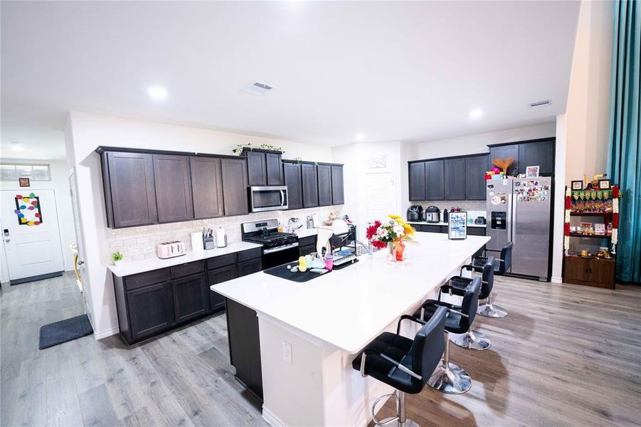 Open-concept kitchen featuring an expansive central island with seating, dark wood-finish cabinetry, stainless steel appliances, and wood-finish flooring Open-concept kitchen featuring an expansive central island with seating, dark wood-finish cabinetry, stainless steel appliances, and wood-finish flooring