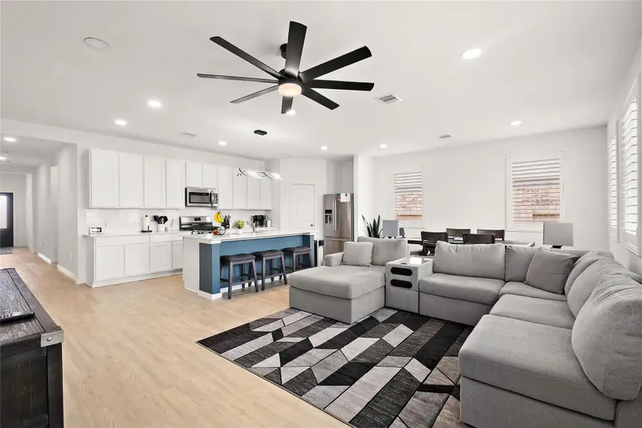 This spacious open-concept living area features a modern kitchen with white cabinetry and stainless steel appliances, a large island with seating, a cozy sectional sofa, and a stylish dining area. Light wood flooring and ample natural light create a warm, inviting atmosphere.