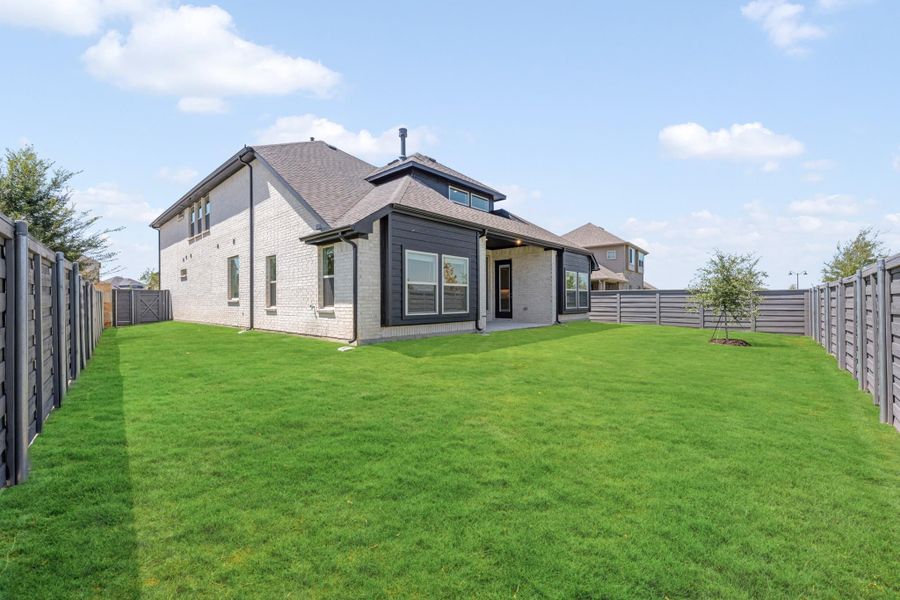 Rear view of two-story brick home with dark trim, covered patio, and large fenced backyard with green lawn