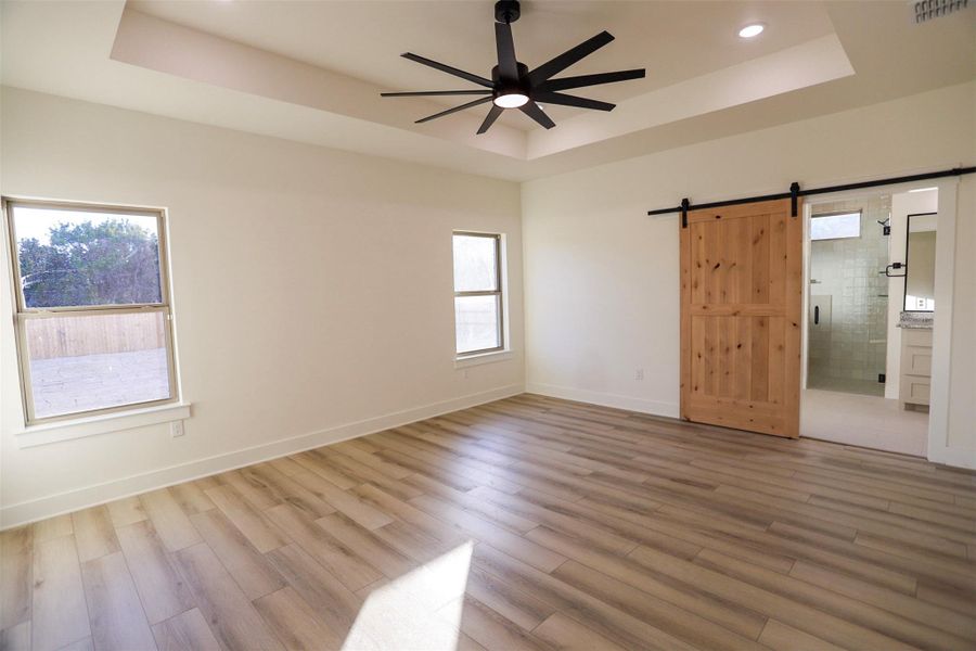 Unfurnished bedroom featuring a barn door, ensuite bath, ceiling fan, light wood-style flooring, and a raised ceiling
