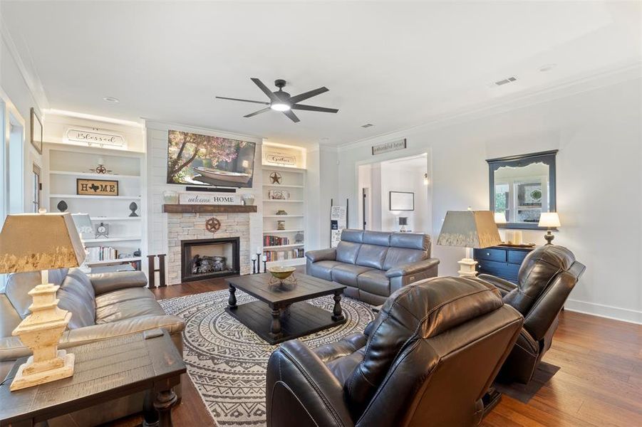 Inviting living room featuring custom built-ins, a stone fireplace with wood mantel, hardwood floors, and abundant natural light, creating a warm and comfortable gathering space.