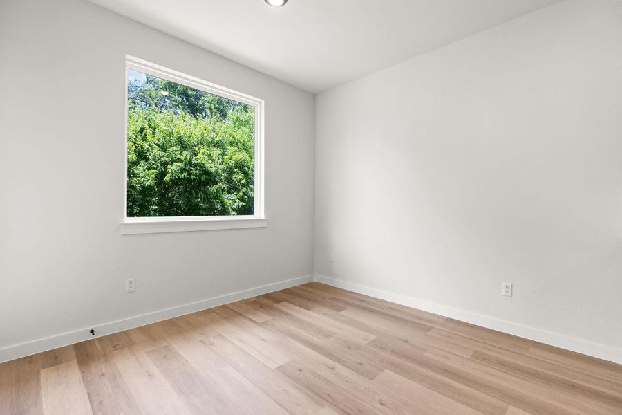 A well-lit bedroom features durable hardwood flooring and a large window overlooking verdant trees.