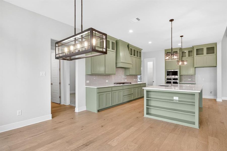 Open-concept kitchen featuring sage green cabinetry, a central island with built-in shelving, light wood-finish flooring, and white countertops Open-concept kitchen featuring sage green cabinetry, a central island with built-in shelving, light wood-finish flooring, and white countertops