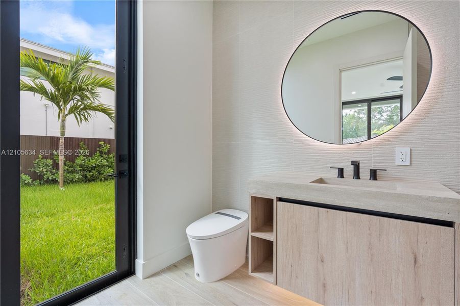 Modern vanity with stone-finish countertop, integrated sink, and wood-finish cabinetry, featuring a circular backlit mirror, wall-mounted matte black faucet, and full-height glass door