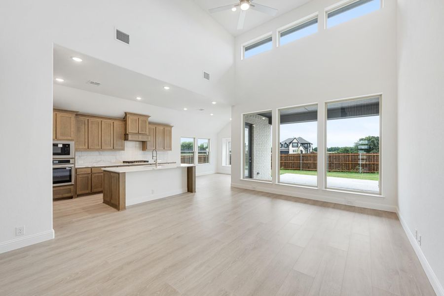 Open living area with high ceilings, light wood floors, and kitchen with island and wood cabinets visible in background Open living area with high ceilings, light wood floors, and kitchen with island and wood cabinets visible in background