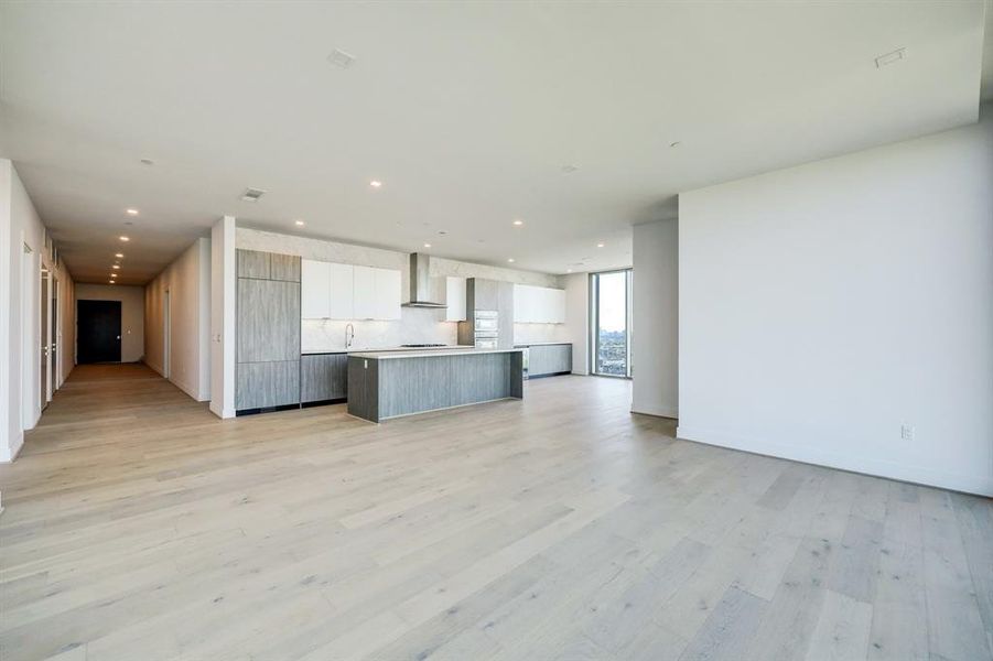 This is a modern, high-rise apartment featuring an open-plan living space with light hardwood floors, a sleek kitchen with a large island, Pedini Italian cabinets, porcelain counter tops and back splash and floor-to-ceiling windows offering expansive city views.