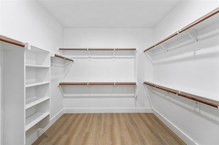 Spacious walk-in closet featuring wood-finish flooring, white walls, built-in shelving, and multiple clothing rods Spacious walk-in closet featuring wood-finish flooring, white walls, built-in shelving, and multiple clothing rods