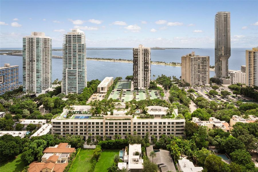Aerial perspective of a multi-story residential complex featuring a rooftop terrace with multiple swimming pools, professional-grade tennis courts, expansive landscaped grounds, and coastal water proximity Aerial perspective of a multi-story residential complex featuring a rooftop terrace with multiple swimming pools, professional-grade tennis courts, expansive landscaped grounds, and coastal water proximity