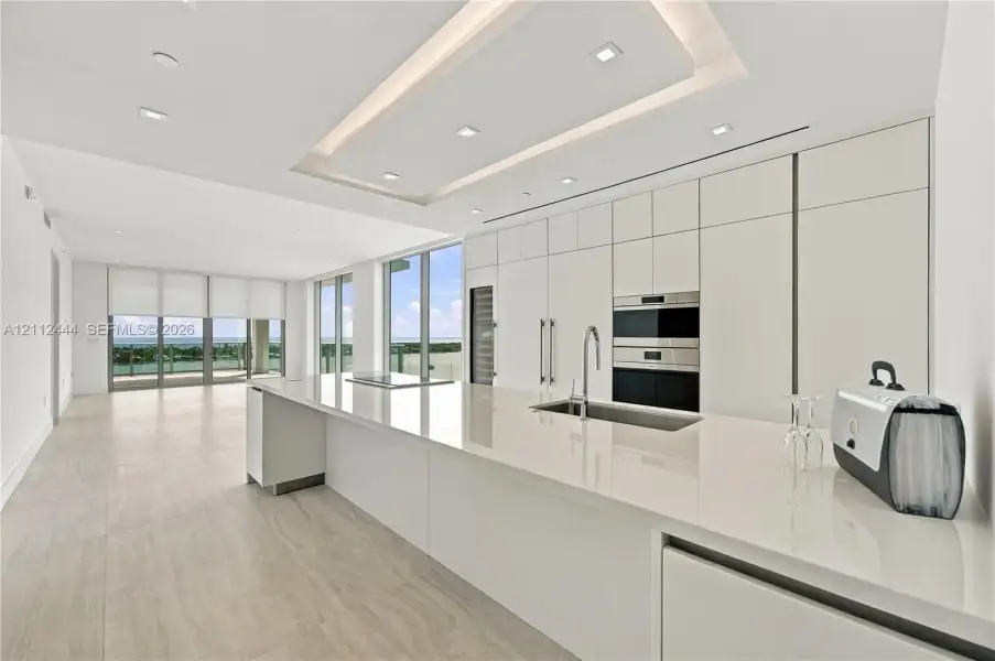 Modern kitchen featuring seamless flat-panel cabinetry, a spacious solid-surface island, built-in stainless steel appliances, recessed tray ceiling lighting, and large-format tile flooring