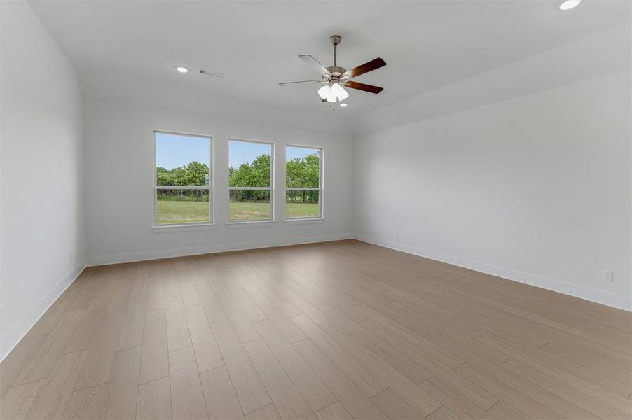 Spacious room featuring light wood-finish flooring, three large windows, a ceiling fan with light kit, recessed lighting, and white baseboards Spacious room featuring light wood-finish flooring, three large windows, a ceiling fan with light kit, recessed lighting, and white baseboards