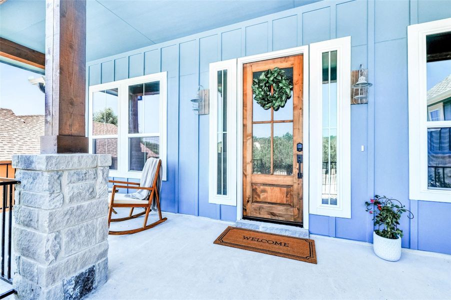 Inviting front porch featuring a natural wood-finish entry door with clear glass panels, board and batten siding, stone-clad pillars, and exterior lighting fixtures