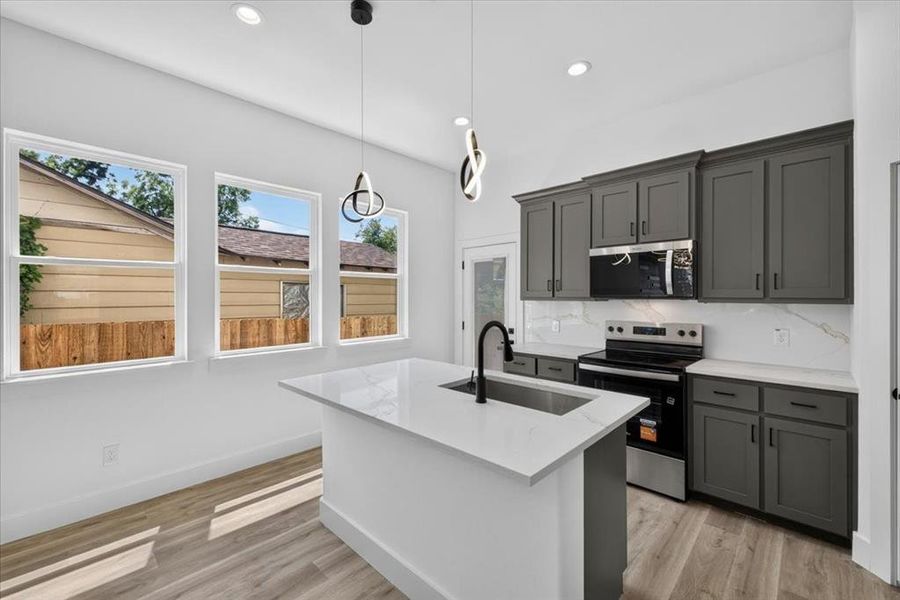 Contemporary kitchen featuring a center island with an undermount sink, quartz countertops, dark gray cabinetry, stainless steel appliances, and wood-finish flooring Contemporary kitchen featuring a center island with an undermount sink, quartz countertops, dark gray cabinetry, stainless steel appliances, and wood-finish flooring