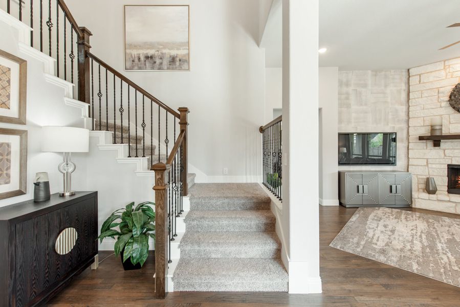 Carpeted staircase with wood and iron railing beside a living room with stone fireplace and hardwood floors