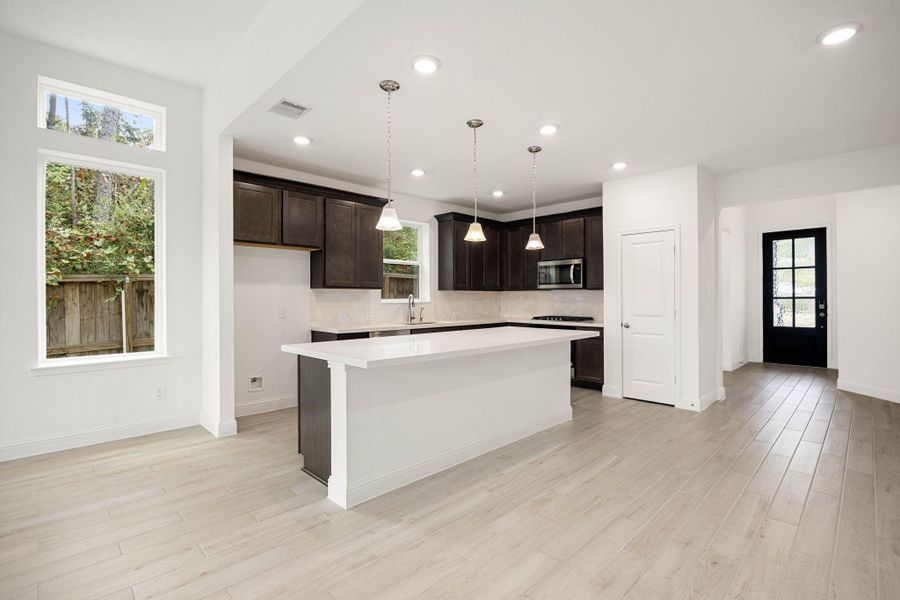 Spacious modern kitchen with sleek dark cabinetry, a large island with pendant lighting, and elegant light wood flooring. Abundant natural light streams through large windows, complementing the open, airy layout.