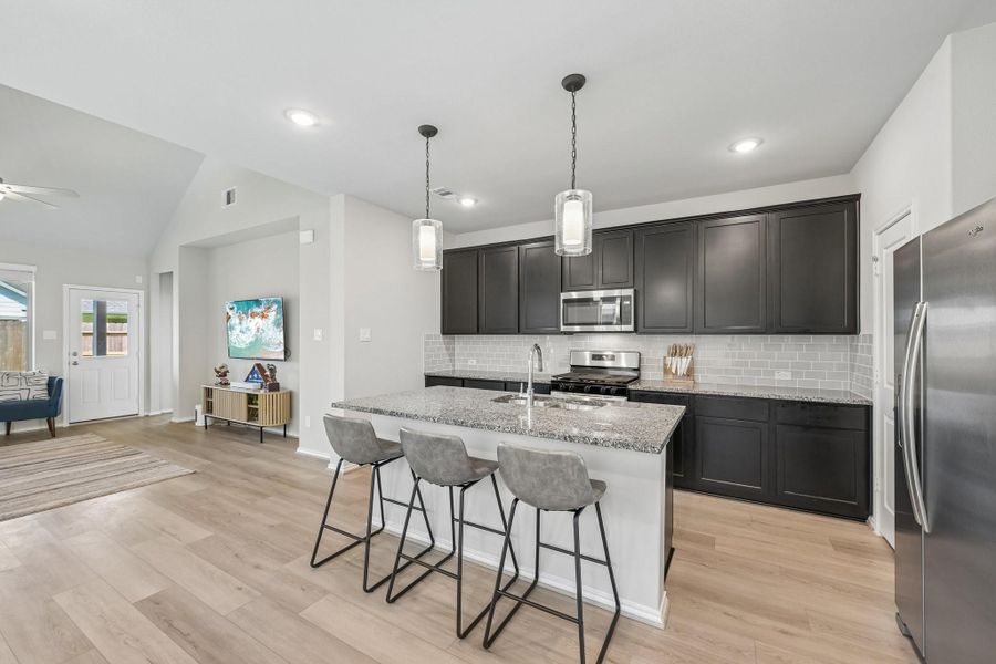 Modern kitchen with sleek dark cabinetry, stainless steel appliances, and a spacious island with seating, seamlessly connecting to an open living area with light wood flooring.