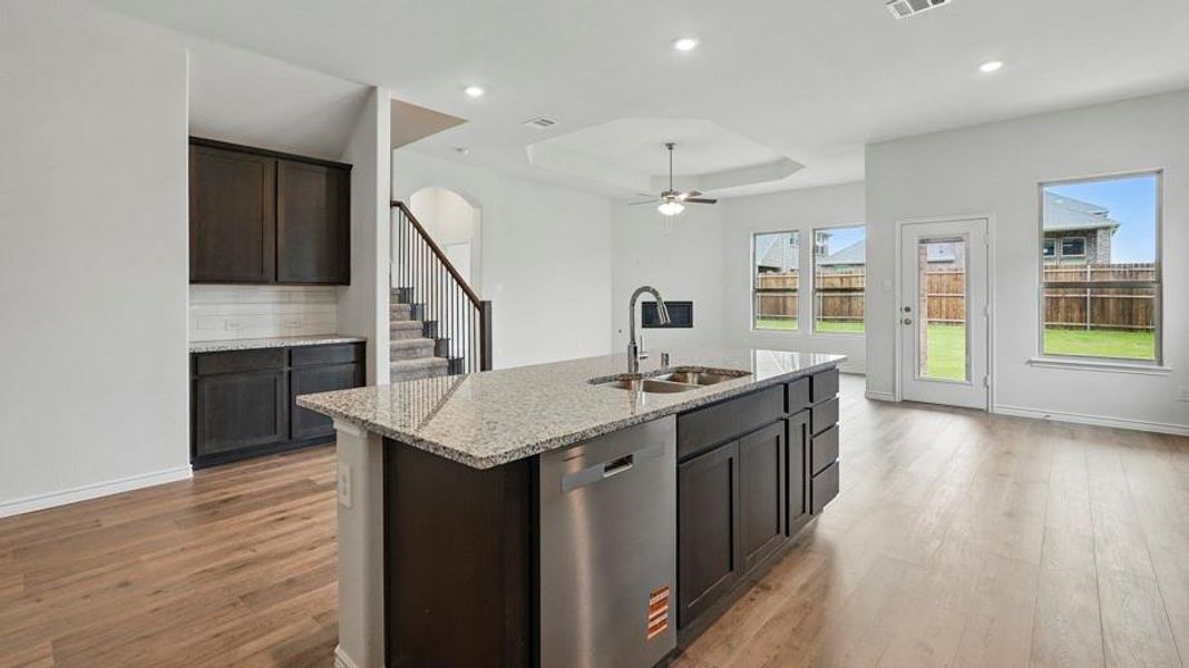 Spacious open-concept living area featuring wood-finish flooring, a kitchen island with stone countertop and integrated sink, stainless steel appliances, and abundant natural light from multiple windows and a glass-paneled door