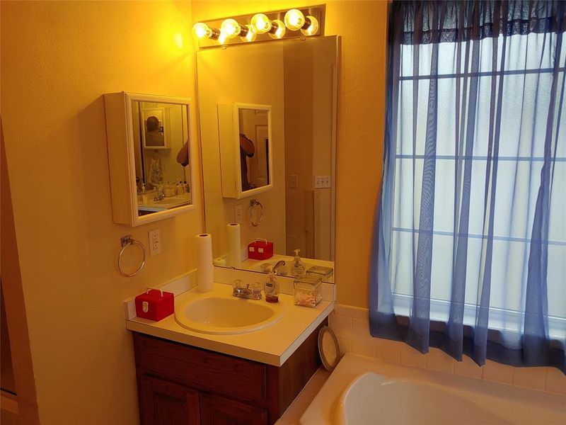 Bathroom featuring a built-in bathtub, a window with vertical mullions, a vanity with a white countertop and integrated sink, a wall-mounted mirror, and overhead strip lighting