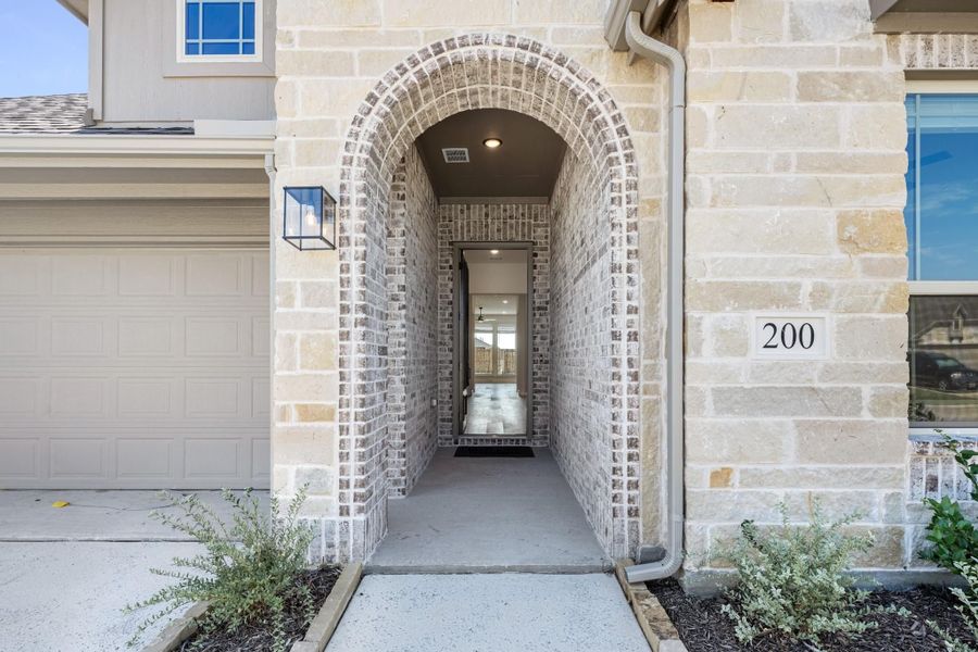 Stone and brick arched entryway leading to front door with wall sconce light and attached garage visible Stone and brick arched entryway leading to front door with wall sconce light and attached garage visible