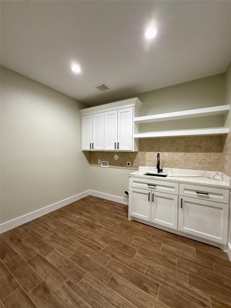 This photo showcases a modern laundry room with sleek white cabinetry, open shelving, and a stylish sink. The room features wood-look tile flooring and recessed lighting, offering a clean and functional space for laundry tasks. This photo showcases a modern laundry room with sleek white cabinetry, open shelving, and a stylish sink. The room features wood-look tile flooring and recessed lighting, offering a clean and functional space for laundry tasks.