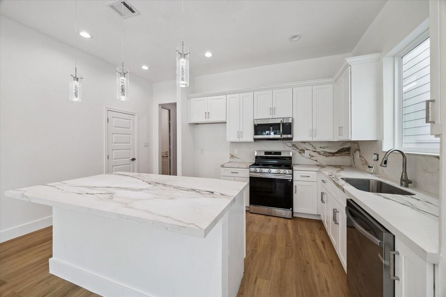 Another view of the kitchen showcases abundant cabinetry, extensive counter space, and a thoughtfully arranged appliance layout. The marble-look backsplash continues seamlessly behind the range and sink for a refined, cohesive finish. Stainless steel appliances, modern hardware, and a large center island give the space a polished look, while natural light and warm flooring create an inviting atmosphere.