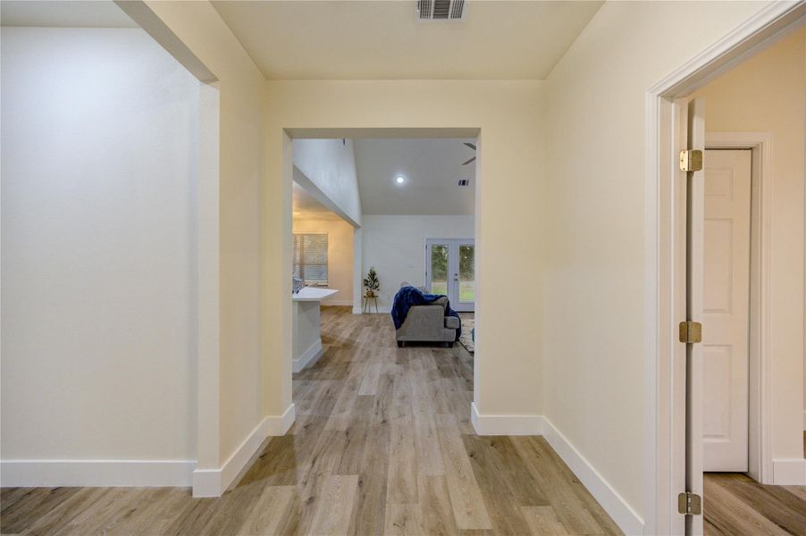 A modern, open hallway with light wood flooring leading into a spacious living area. The living room features high ceilings, recessed lighting, and large windows with a view of the backyard, creating a bright and inviting space.