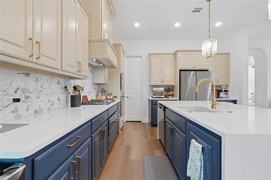Contemporary kitchen featuring a large central island with a gold-tone faucet, white countertops, two-tone cabinetry, wood-finish flooring, and a patterned backsplash Contemporary kitchen featuring a large central island with a gold-tone faucet, white countertops, two-tone cabinetry, wood-finish flooring, and a patterned backsplash