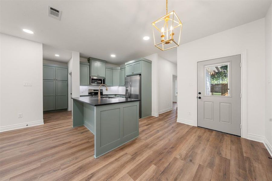 Kitchen featuring dark countertops, a kitchen island with sink, green cabinets, decorative light fixtures, and a chandelier Kitchen featuring dark countertops, a kitchen island with sink, green cabinets, decorative light fixtures, and a chandelier