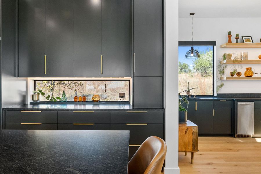Modern kitchen featuring dark cabinetry with gold-tone hardware, a horizontal window backsplash, and dark stone countertops