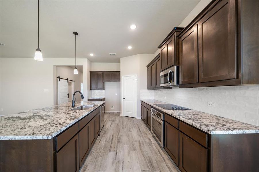 Kitchen with a barn door, backsplash, light wood-type flooring, dark brown cabinetry, and light stone counters Kitchen with a barn door, backsplash, light wood-type flooring, dark brown cabinetry, and light stone counters