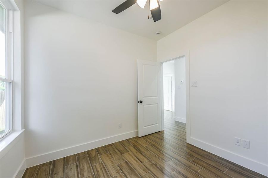 This is a bright, empty room featuring wood flooring, a large window for natural light, and a ceiling fan. It includes a standard door leading to another room.