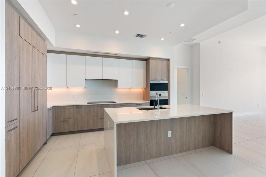 Kitchen featuring two-tone cabinetry with wood-finish and matte white finishes, a large central island with a solid-surface countertop, integrated wall-mounted double ovens, recessed lighting, and tile flooring