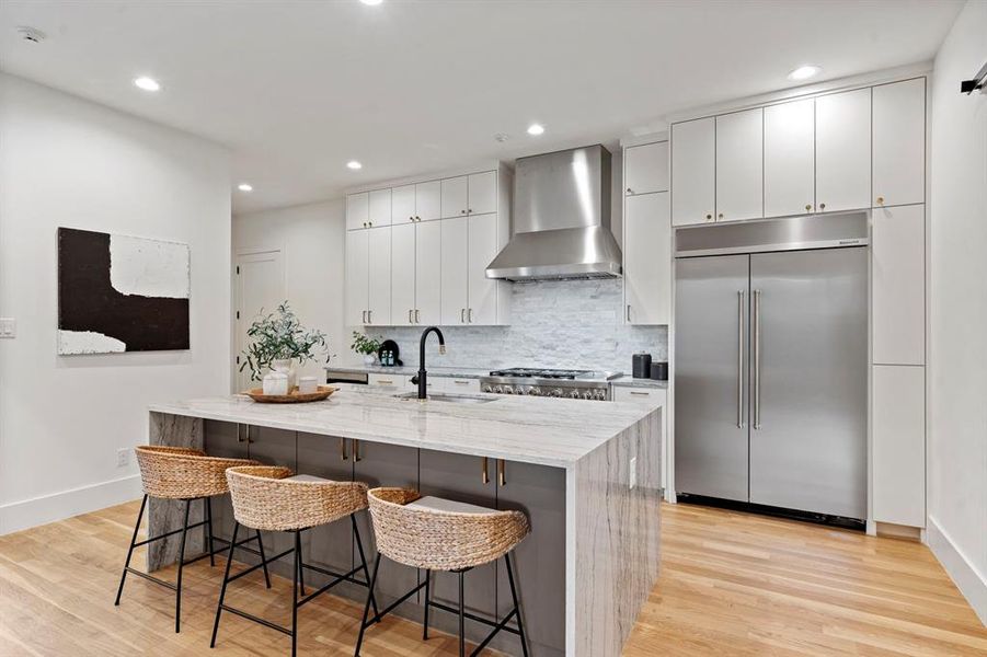 Kitchen featuring built in fridge, wall chimney exhaust hood, decorative backsplash, a kitchen breakfast bar, and light wood-type flooring Kitchen featuring built in fridge, wall chimney exhaust hood, decorative backsplash, a kitchen breakfast bar, and light wood-type flooring