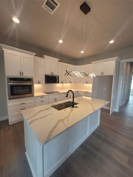 Kitchen featuring light stone counters, appliances with stainless steel finishes, white cabinets, hanging light fixtures, and an island with sink
