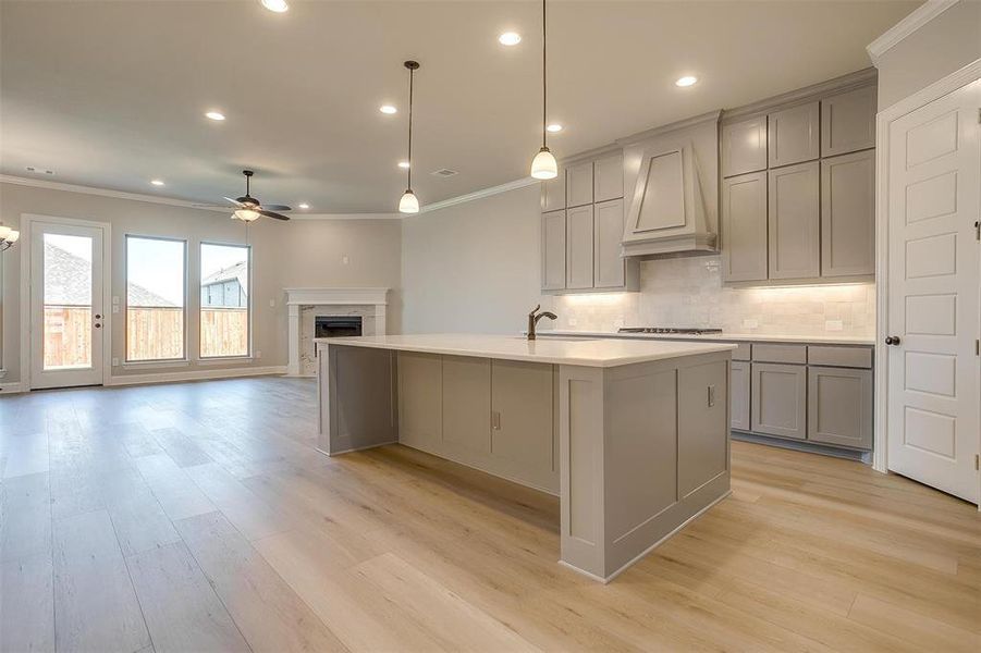 Kitchen with custom range hood, ornamental molding, tasteful backsplash, light wood-type flooring, and a kitchen island with sink