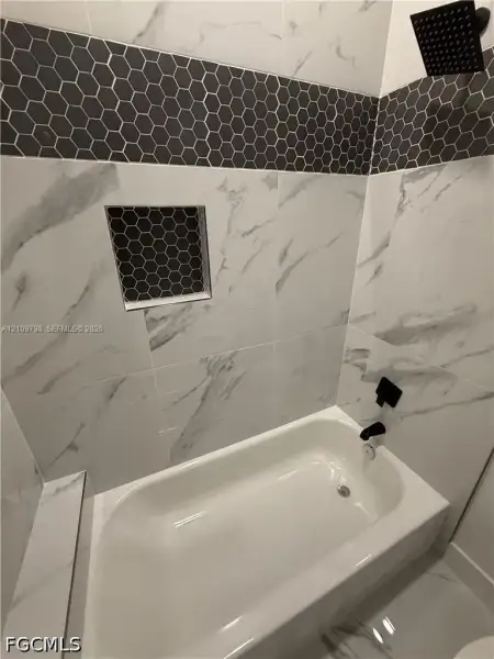 Bathtub surround featuring large-format white stone-finish tile, a charcoal hexagonal tile accent border, an integrated recessed niche, and matte black plumbing fixtures including a rainfall shower head