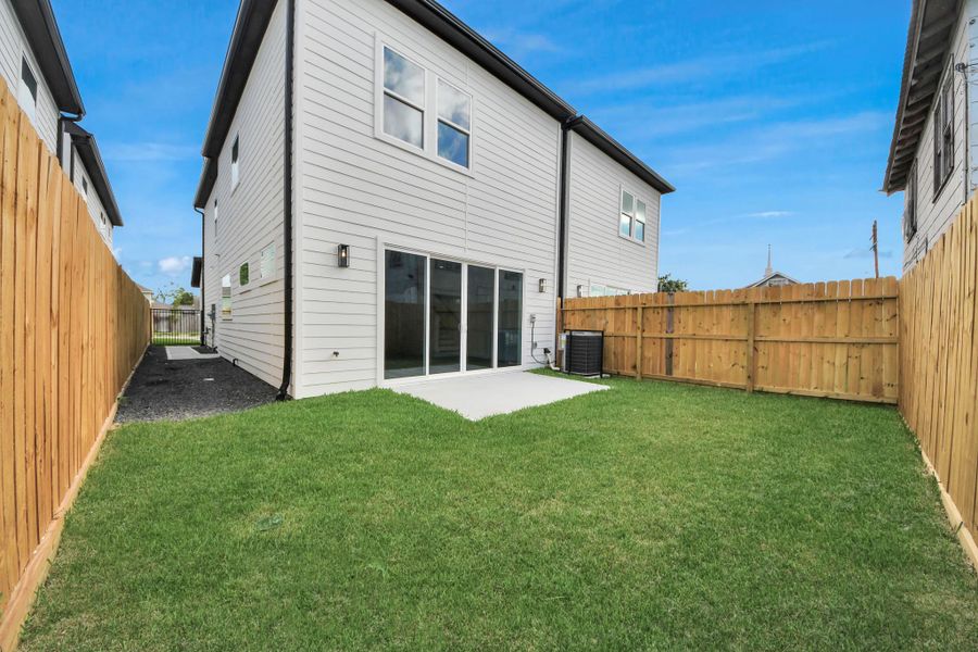 This photo shows the backyard of a modern two-story home with a well-maintained lawn and a wooden fence for privacy. It features a small concrete patio area with sliding glass doors leading into the house, offering a cozy outdoor space for relaxation.