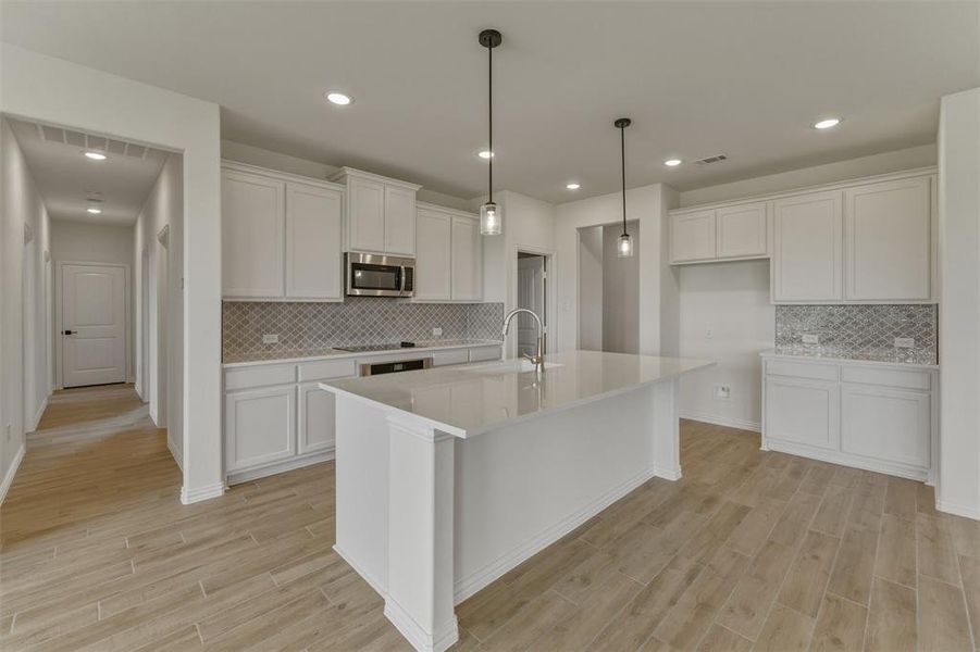 Modern kitchen featuring a large central island with a sink and pendant lighting, white cabinetry, integrated stainless steel microwave, and wood-finish flooring Modern kitchen featuring a large central island with a sink and pendant lighting, white cabinetry, integrated stainless steel microwave, and wood-finish flooring