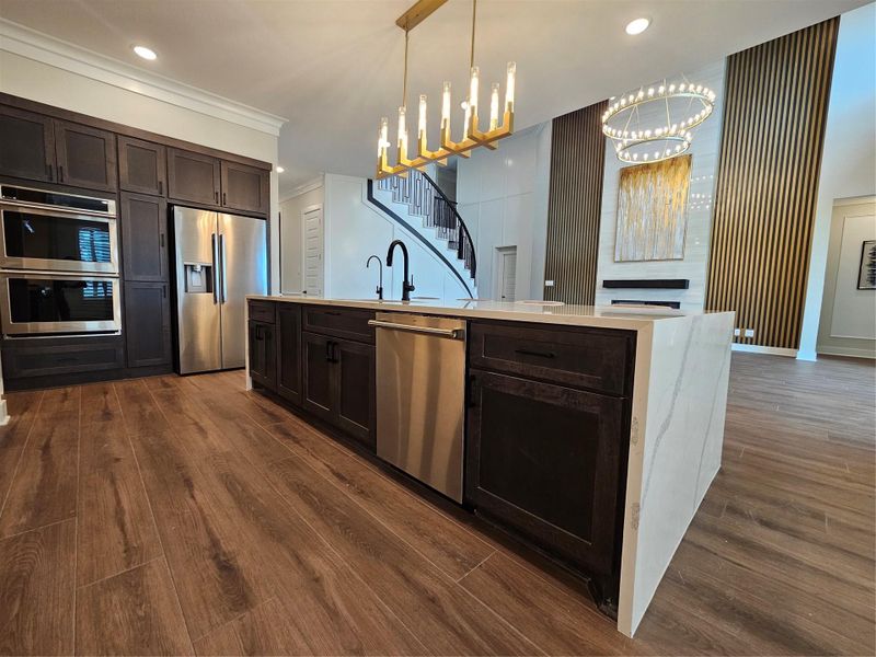 This modern kitchen features a large island with a marble countertop, dark wood cabinetry, and stainless steel appliances. The open layout flows into a stylish living area with elegant lighting, wood flooring, and a striking staircase.