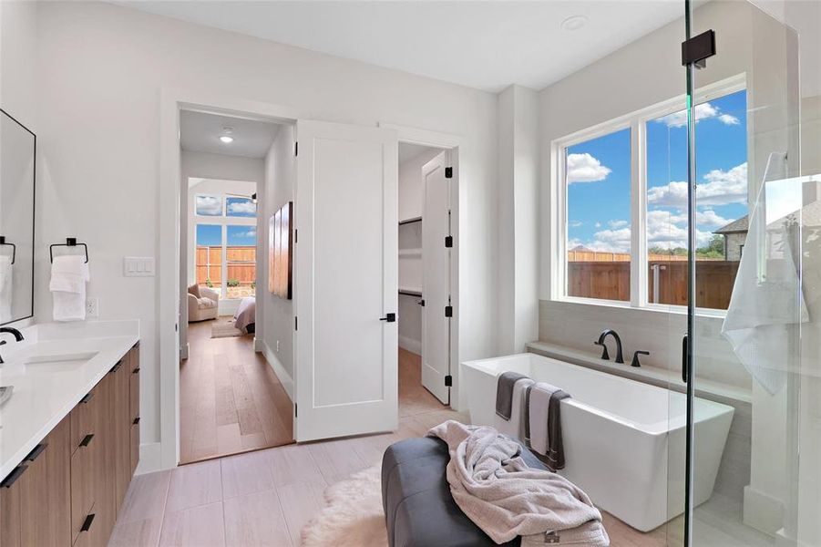 Contemporary bathroom featuring a freestanding tub, large window, glass-enclosed shower, dual vanity with wood-finish cabinetry, and light wood-look flooring