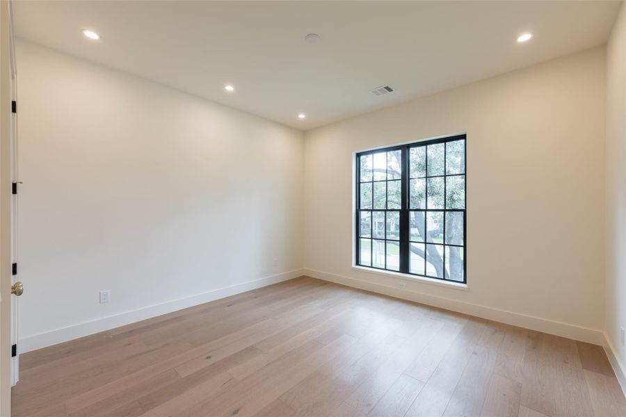 This secondary bedroom features wide-plank white oak floors and large black-trimmed windows that frame views of mature trees and the front lawn. Clean lines and recessed lighting offer a versatile backdrop for any design style. This secondary bedroom features wide-plank white oak floors and large black-trimmed windows that frame views of mature trees and the front lawn. Clean lines and recessed lighting offer a versatile backdrop for any design style.