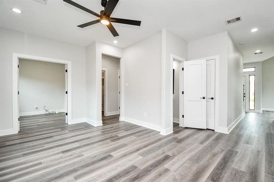 This is a view from kitchen of this bright, spacious family room with modern grey wood flooring, crisp white walls, and a contemporary black ceiling fan. The space features multiple doorways leading to adjacent study or office and a large under stairs  closet for storage. The room benefits from recessed lighting and has a clean, airy feel.