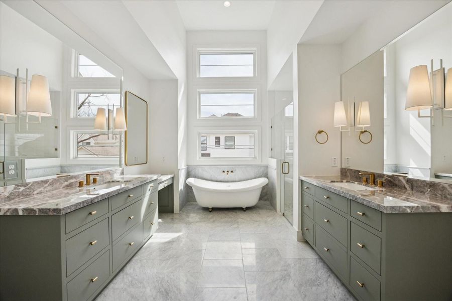 Spa inspired primary bath stuns with dual sage vanities topped in dramatic marble and accented by warm brass fixtures. Freestanding soaking tub framed by stacked picture windows creates a light filled focal point, while a glass enclosed shower completes the retreat experience.