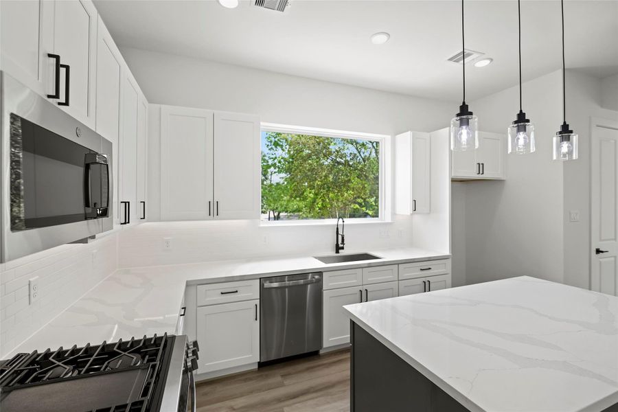 This modern kitchen features sleek white cabinetry, stainless steel appliances, and a large window providing ample natural light. The marble-like quartz countertops and stylish pendant lights over the island create an elegant and functional space.