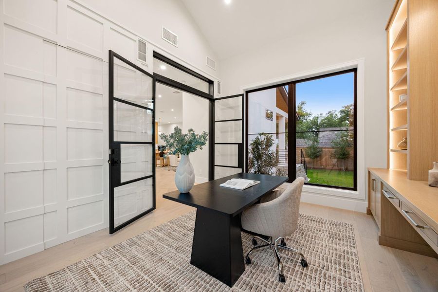 A stylish home office designed for productivity and comfort, featuring large windows that bring in natural light and views of the outdoors. Clean architectural details and modern finishes create a refined workspace. The highlight of this room is the glass-paneled doors, adding a sophisticated design element while maintaining an open and connected feel to the rest of the home.