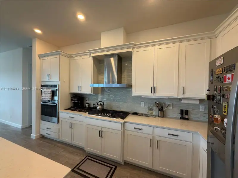 Kitchen featuring white shaker-style cabinetry, a stainless steel chimney-style range hood, integrated double ovens, and a neutral-toned tile backsplash