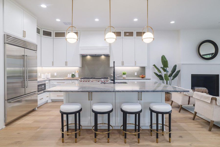 Open-concept kitchen featuring an oversized island with seating, stainless steel appliances, white cabinetry with gold hardware, a tile backsplash, and wood-finish flooring