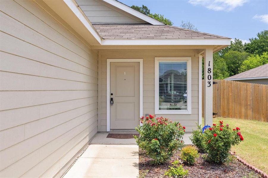 Inviting home entrance featuring horizontal siding, an entryway with a covered porch, a privacy fence, and manicured landscaping with flowering shrubs