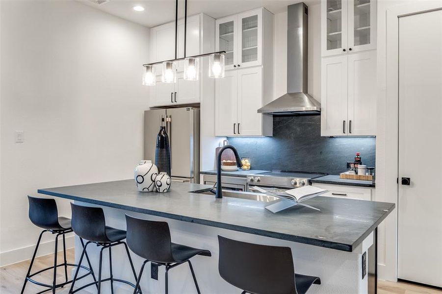 Kitchen featuring appliances with stainless steel finishes, hanging light fixtures, white cabinets, wall chimney exhaust hood, and a kitchen island with sink