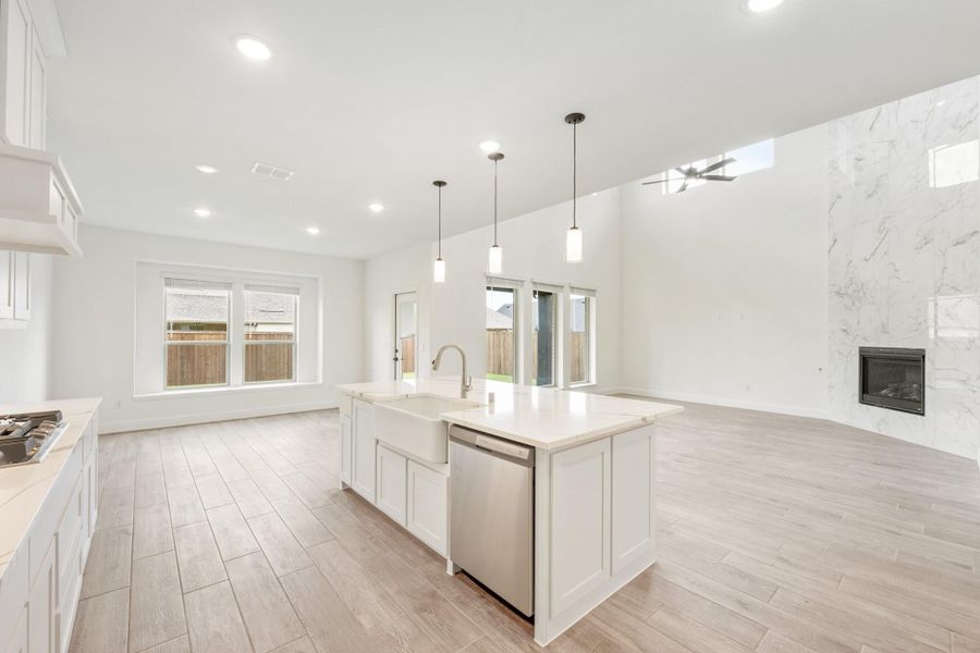 Open-concept kitchen with white island, farmhouse sink, and pendant lights, flowing into living area with marble fireplace